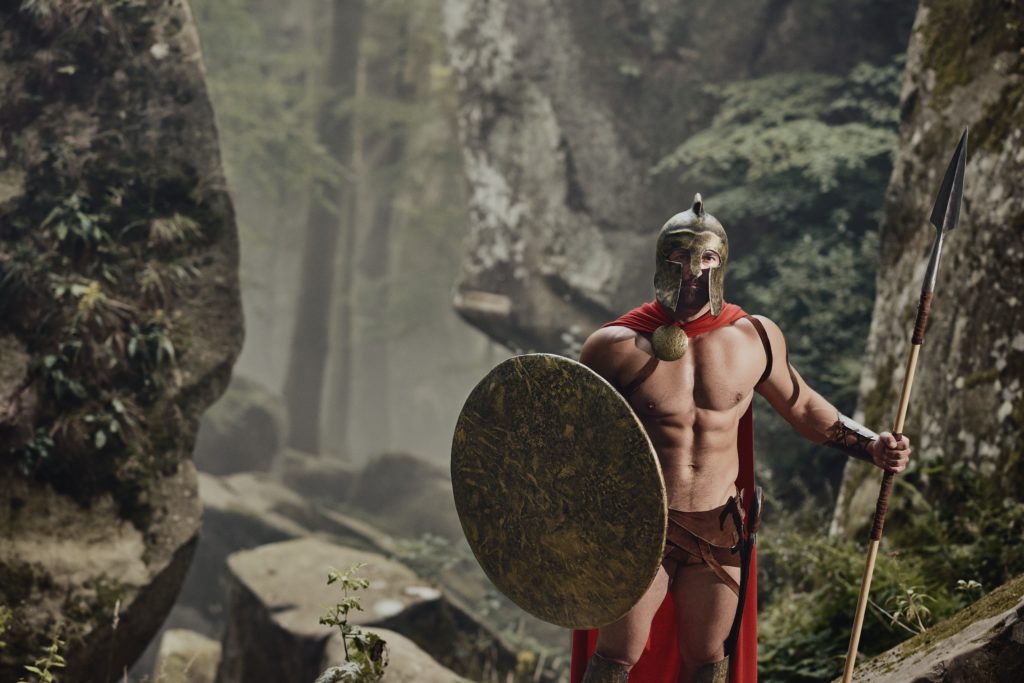 Young muscular man holding shield and spear posing in armor of ancient gladiator looking at camera on background of woods. Spartan.